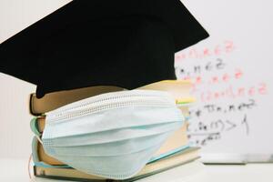 The black square hat of the master lies on the textbooks stacked in a column and decorated with a protective medical mask. Education concept photo