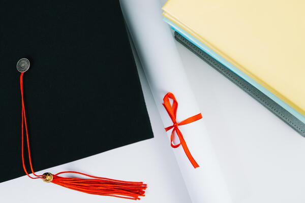 A black square graduate hat with a red tassel lies on a diploma and a stack of books. Education concept