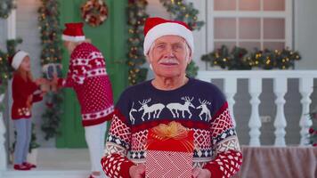 Portrait of grandfather man presenting gift box smiling near decorated Christmas house with family video