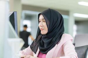joven musulmán mujer de negocios en un oficina, joven Arábica mujer en un Pañuelo utilizando un ordenador personal computadora foto