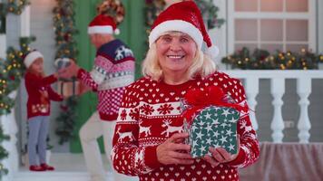 Portrait of grandmother woman presenting gift box smiling near decorated Christmas house with family video