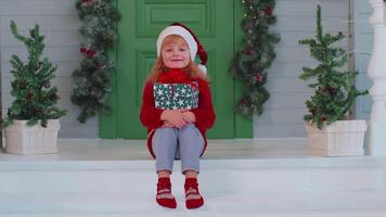 Cheerful smiling toddler child girl kid sitting at decorated house porch holding one Christmas box video