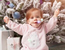 Small girl in the pink pajama sitting near the christmas tree playing with the garland photo