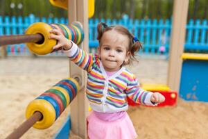 a little girl with two tails is dressed in a striped colorful jacket is playing in the sandbox on the playground photo