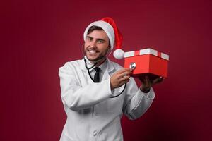 Young handsome doctor in white uniforme and Santa Claus hat standing in studio on red background smile and finger in camera photo