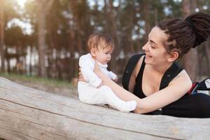 A beautiful smiling young brunette woman with long dreadlock hair hold a pretty baby. Mother looks tenderly at the child. They are in the green summer forest. photo