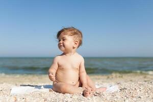 pequeño niño sentado en un toalla por el mar desnudo alegre y contento foto