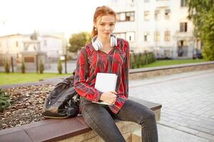 young beautiful student wearing a checkered shirt headphones and a backpack resting on the street between classes photo