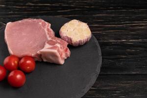 Top view of one pieces raw pork chop steaks with cherry tomatoes and garlic on a black stone cutting board. photo