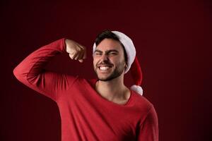 Young handsome caucasian guy in a red sweater and Santa hats stands on red background in studio and showing biceps power photo