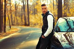 un hermoso joven caucásico hombre vistiendo estilo negro chaleco en pie cerca negro coche en naturaleza. foto