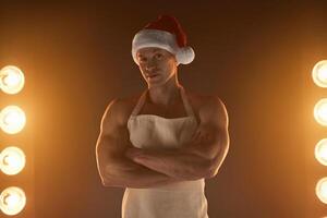 caliente Navidad postre. muscular hombre vistiendo delantal y Papa Noel sombrero doblada brazos y sonriente a cámara, lamparas iluminación en antecedentes foto