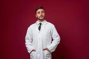 Young handsome modern doctor in a white medical gown stands in the studio on a red background. Student trainee of a medical university. photo