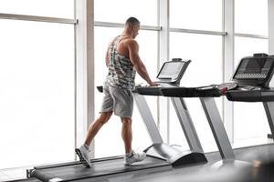 Muscular athletic bodybuilder fitness model running treadmill gym near big window photo
