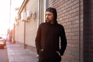 Handsome young unshaven man black hoodie and bandana stands backyard near brick walls summer day. photo