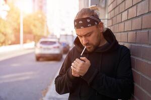 Handsome young unshaven man black hoodie and bandana stands backyard near brick walls summer day. photo