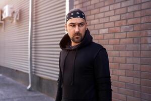 Handsome young unshaven man black hoodie and bandana stands backyard near brick walls summer day. photo