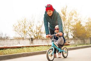 A young Caucasian mother teaches a little daughter to ride a runbike on a warm autumn day. photo