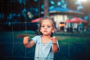 the little girl holds on to the fence with both hands and looks into the camera through the grid photo