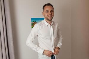 hermoso hombre poniendo en camisa en pie cerca ventana a su habitación en Mañana foto