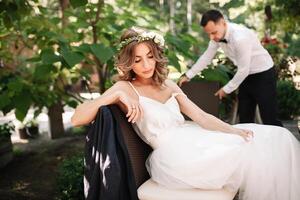 A beautiful bride in a white wedding dress and wreath sits on a chair next to the groom, resting and preparing for a happy family life photo