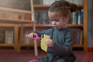 linda pequeño caucásico niña es jugando con tijeras y de colores papel mientras sentado en el piso a hogar. foto