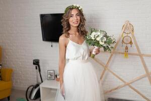 The bride in a beautiful wedding dress with a long curly hair stands in the bedroom and looks at the camera. Bride indoors with white flowers bouquet and tender wreath hairstyle photo