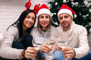 contento amigos celebrando nuevo año en hogar interior en Navidad sombreros sentado cerca un Navidad árbol con lentes de vino foto