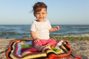 contento bebé sonriente y ondulación mano, sentado en blanco arenoso tropical playa en el alfombra foto