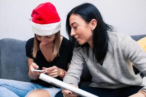 dos joven mujer son preparando para el nuevo año mirando en móvil teléfono formas a paquete regalos. nuevo año celebracion foto