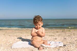 pequeño niño sentado en un toalla por el mar desnudo alegre y contento foto