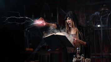 Halloween concept. Witch dressed black hood with dreadlocks standing dark dungeon room use magic book for conjuring magic spell photo