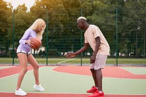 Active multiethnic couple playing basketball on outdoor sport court, black guy teaching his blonde girlfriend to play streetball photo