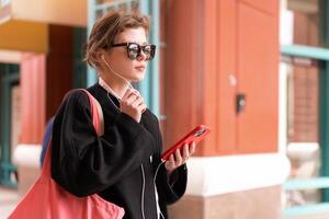 Portrait serious nice woman wearing black coat listening music with headphones and using smartphone. Beautiful caucasian female have phone conversation with earphones and mobile phone outdoor. photo