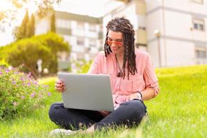 Photo of european woman 25s sitting on green grass in park with legs crossed during summer day while using laptop. Caucasian female hipster with dreadlocks and pink sunglasses on use laptop