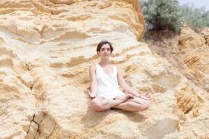 un hermosa joven niña con corto pelo es vestido en pantalones cortos y un blanco jersey es practicando yoga en el antecedentes de rocas actitud de el loto. el concepto de calma y concentración foto