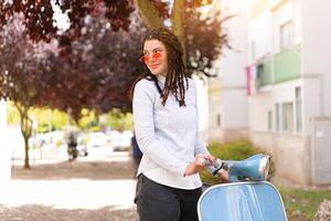 Hipster girl in pink sunglasses with dreadlocks standing near vintage motor scooter on city street photo