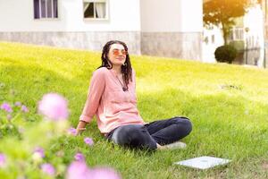 Photo of european woman 25s sitting on green grass in park with legs crossed during summer day while using laptop. Caucasian female hipster with dreadlocks and pink sunglasses on use laptop