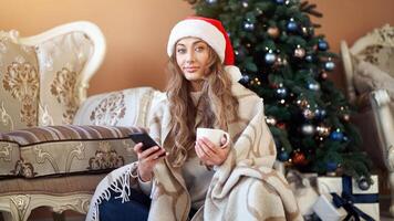 Christmas. Woman dressed white sweater Santa hat on the floor near christmas tree with smartpone and cup of coffee photo