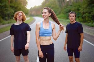 A group of three people athletes one girl and two men standing on asphalt road in pine forest at summer before jogging photo