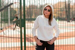 Outdoor close up portrait of young beautiful woman with long hair in sunglasses, dressed in a white sweater, near the sportsground photo