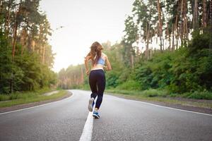 Beautiful caucasian young girl athlete runs sunny summer day on asphalt road in the pine forest view from behind. photo