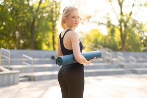 Sporty Slim Woman Sportive Clothes Black Leggings and Top Standing near concrete Stairs Holding Yoga Mat Resting Between Exercises. Sport and Fitness Concept photo