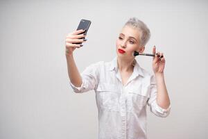 Studio portrait of beautiful trendy girl with short hair and make up holding arm on waist and taking selfie via cell phone over white isolated background photo