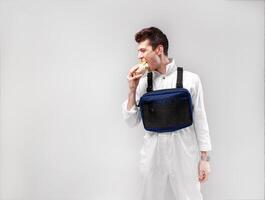 Young stylish male worker with chest rig bag is eating a tasty juicy burger on a white background photo