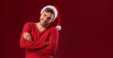 joven hermoso caucásico chico en rojo suéter y Papa Noel sombreros soportes en rojo antecedentes en estudio y dientes sonriente foto