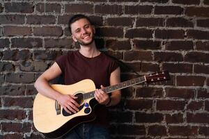 Man with acoustic guitar against brick wall playing music singing songs enjoy life Medium shoot photo