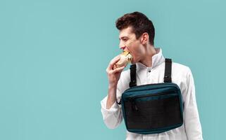 Young stylish male worker with chest rig bag is eating a tasty juicy burger on a white background photo