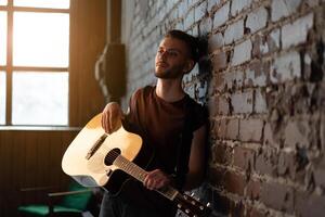 Man with acoustic guitar leaning against brick wall playing music singing songs enjoy life Medium shoot photo