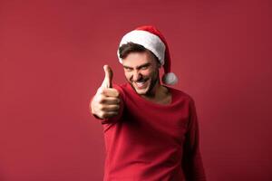 Young handsome caucasian guy in a red sweater and Santa hats stands on red background in studio and showing thumbs up. photo